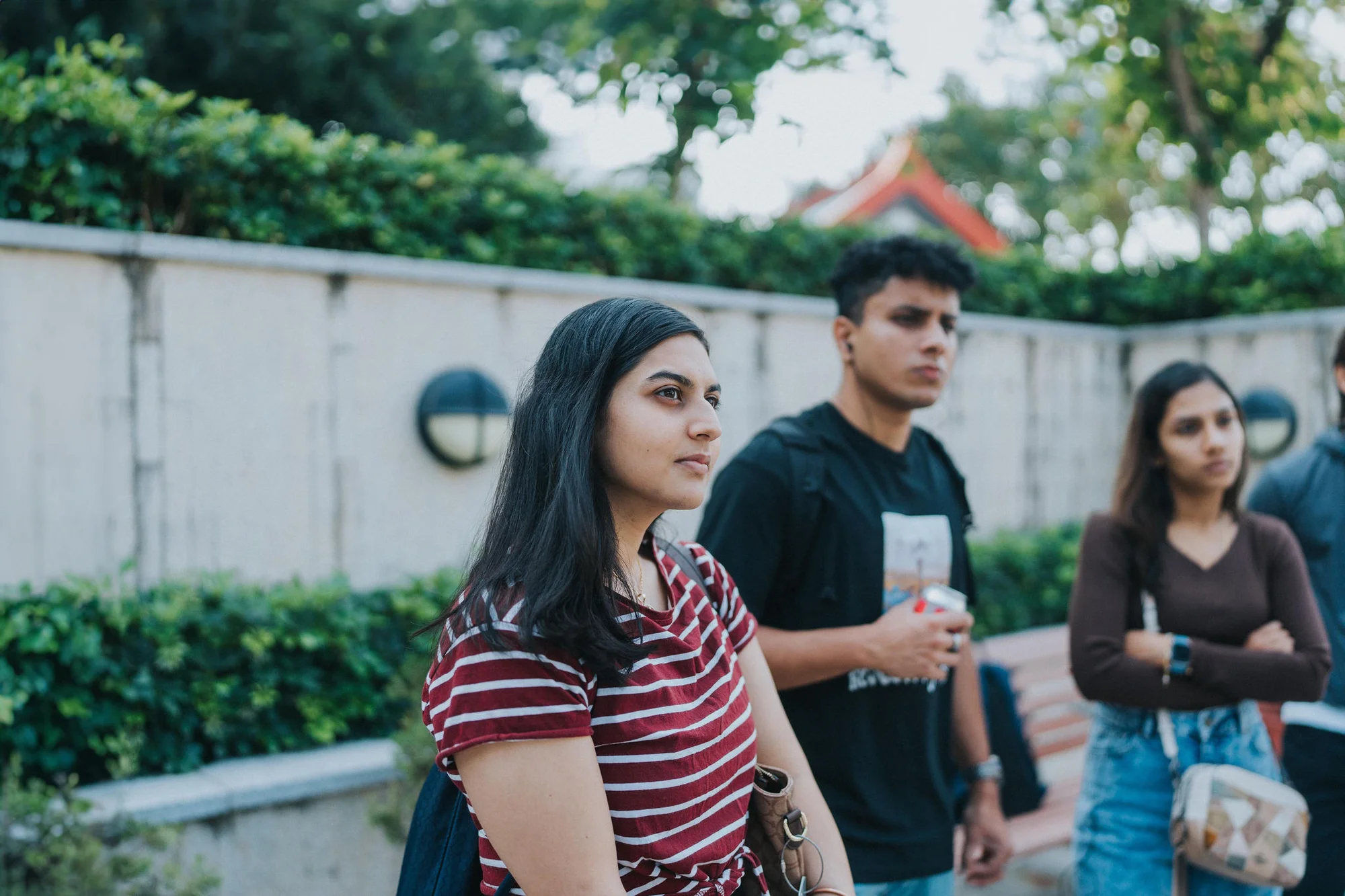 A close-up shot of a woman in a red striped shirt, standing confidently with her arms crossed and a slight smile on her face. Beside her is a group of young adults (tourists) enjoying each other's company on a warm summer day outdoors. There is a man with short hair holding a can and a young woman with long black hair standing next to a tree gazing off into the distance.